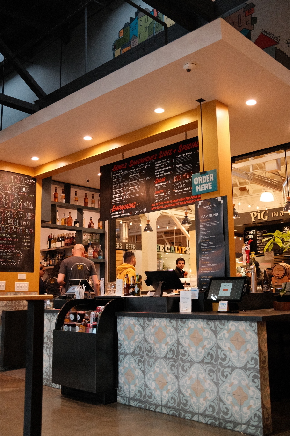 Interior of a restaurant with a menu board displaying various food and drink options, a bar counter with bottles, and a few people behind the counter.